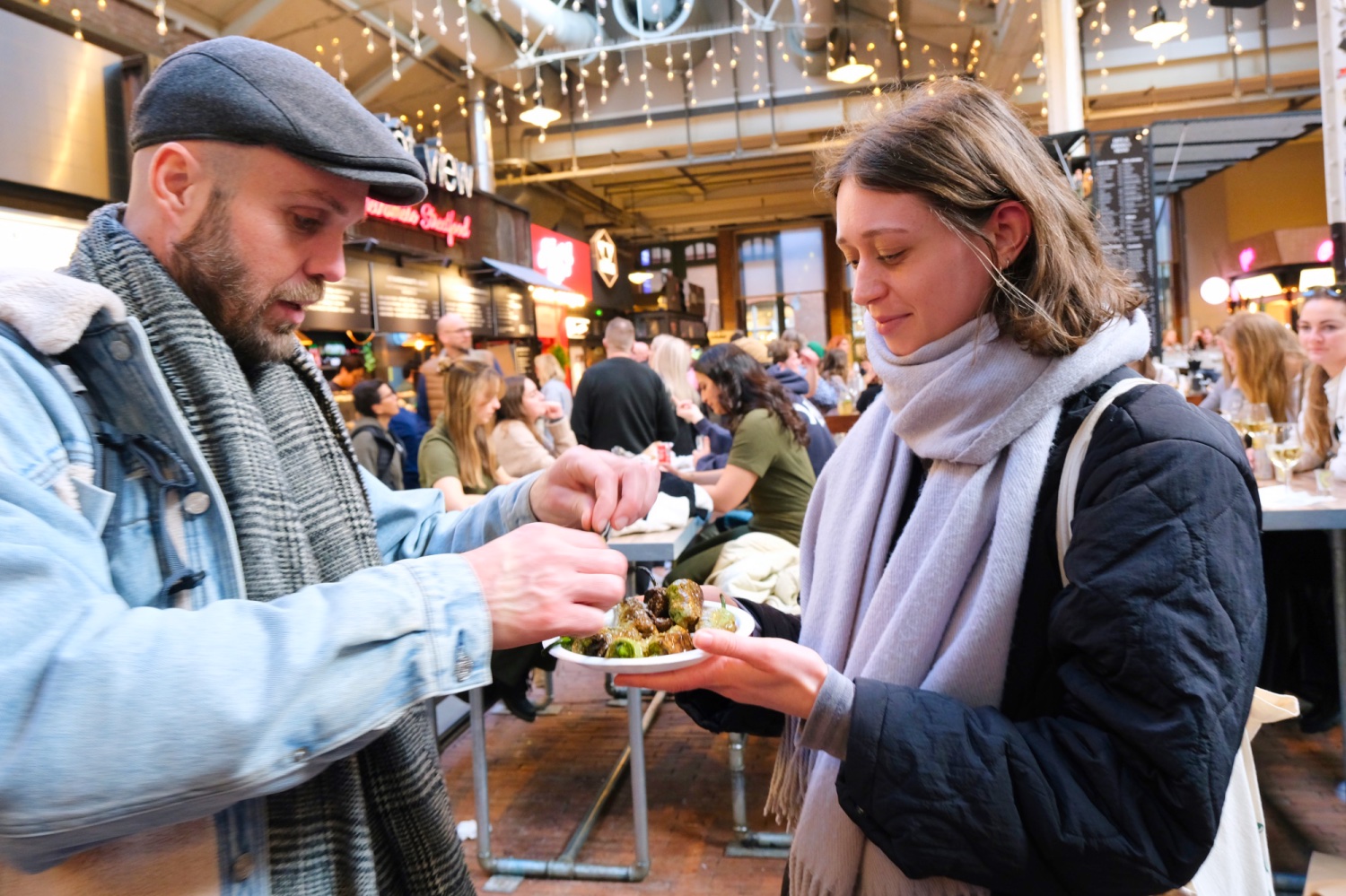 In de Foodhallen in Amsterdam staan twee personen. De ene heeft een bordje van padron pepers vast en de ander pakt een peper vast.