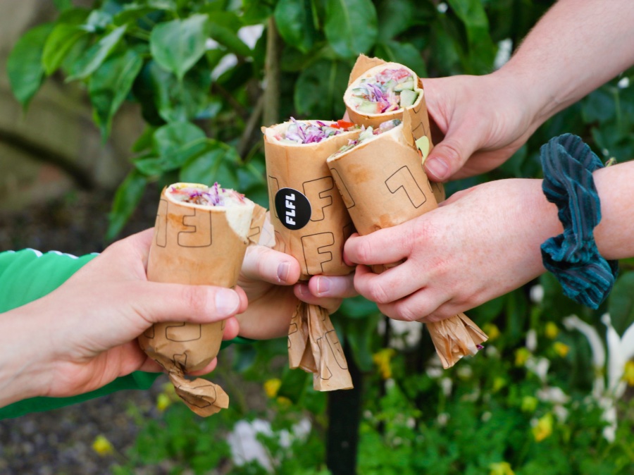 Vier handen hebben allemaal een halve falafel rol vast en ze 'proosten' hiermee met elkaar