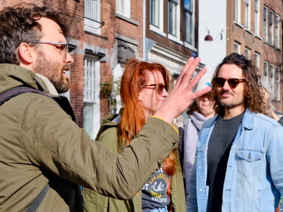 Voor een aantal Amsterdamse huizen zie je een groep mensen. Een gids is iets aan het vertellen en de rest van de mensen luistert aandachtelijk