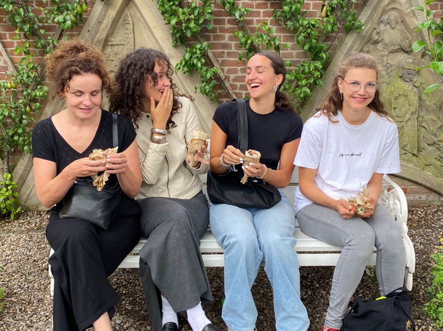 Op een bankje zie je vier mensen naast elkaar zitten met een wrap met falafel in hun hand. Iedereen lacht breed