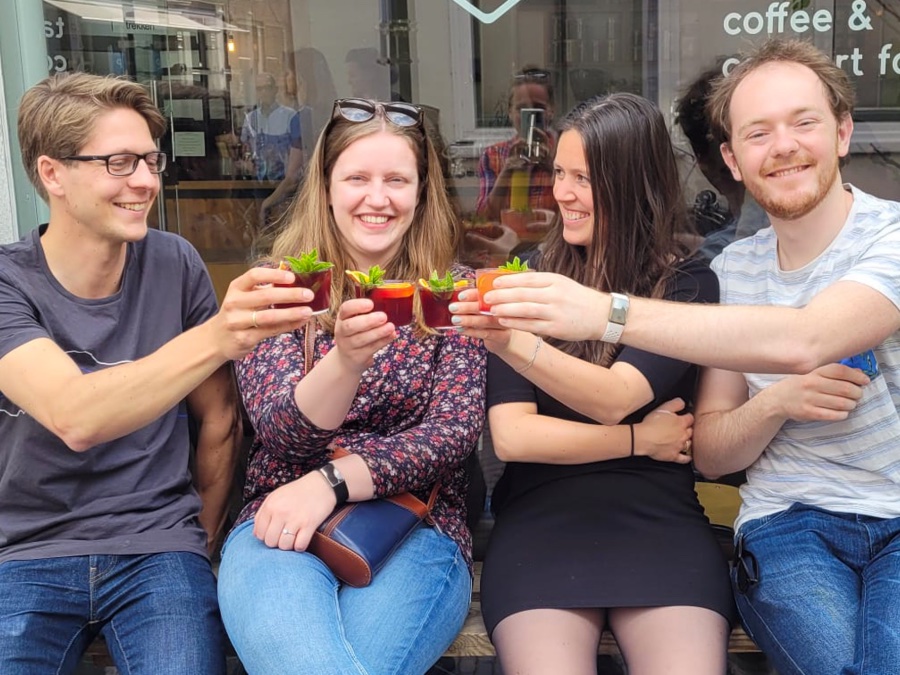 Op een bankje voor een restaurant zitten vier mensen naast elkaar, ze proosten met een drankje in hun hand