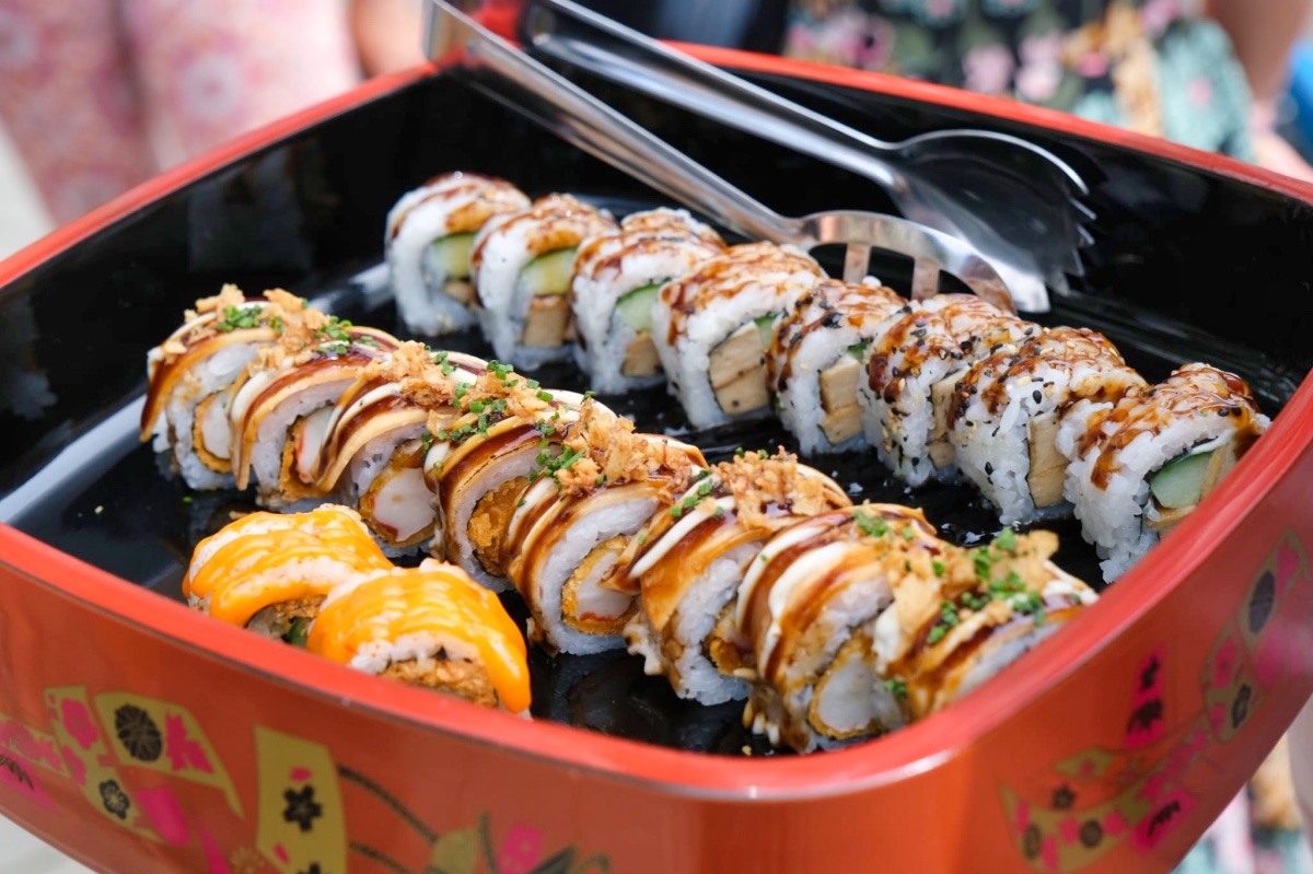 In een rode Japanse bak zie je allemaal mooigesneden sushirollen. Er bij een tang waarmee je de sushi kan oppakken.