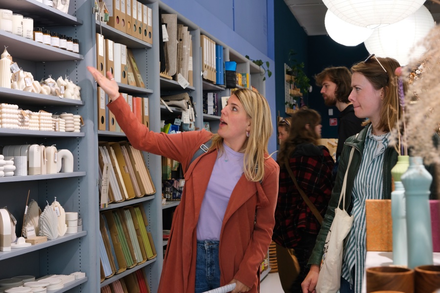 In een winkel met witte kaarsen staan allemaal klanten. Een gids met een oranje jas en paars shirt wijst naar de kaarsen en vertelt iets terwijl de rest luistert naar het verhaal.