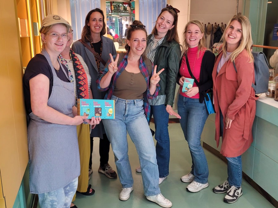 In een winkel staan zeven personen. Ze kijken allemaal lachend de camera in. Het is een kleurrijk geheel. Een iemand die links voor straat houdt een doosje vast met koeken.