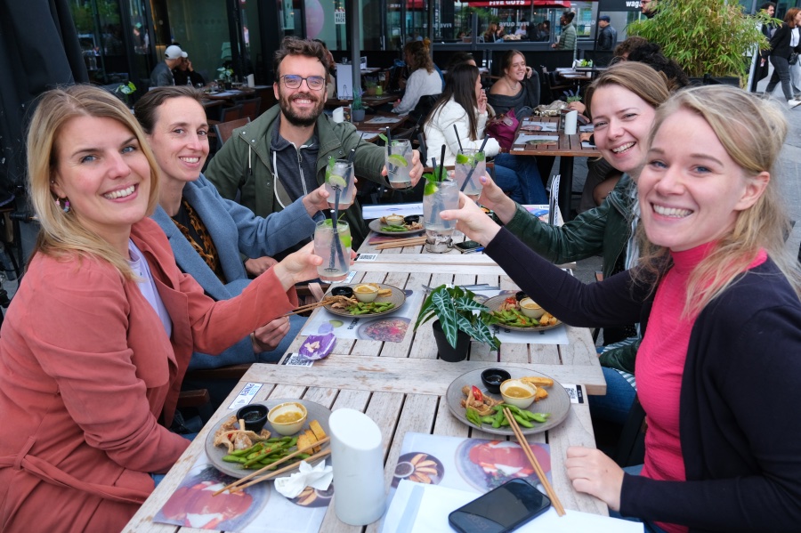Om een tafel zie je vijf personen. Zij proosten met elkaar met een drankje met een limoentje en een rietje. Iedereen kijkt in de camera. Voor zich hebben ze allemaal een bordje met eten