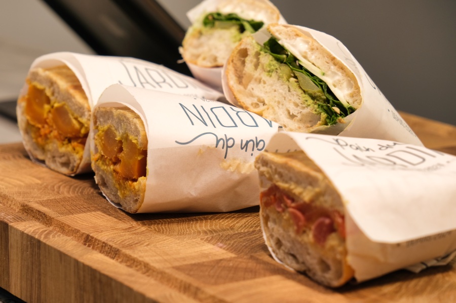 Op een houten plank zie je vijf broodjes in een witte verpakking. Het zijn broodjes die door de midden zijn gesneden met daarop groenten en een gele saus