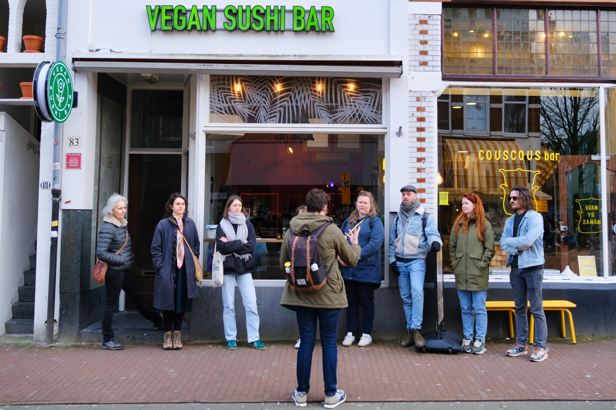 Een groep mensen staat voor de Vegan Sushi Bar in Amsterdam en een gids vertelt een verhaal.
